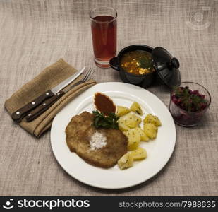 Business lunch : Petite Wiener schnitzel with boiled potatoes and ketchup. Served on a white porcelain plate with fork and knife on a wooden background. Beet salad and tomato, red pepper soup, sauce with olive oil, rosemary and smoked paprika with fork and a glass of juice and knife on a wooden background.