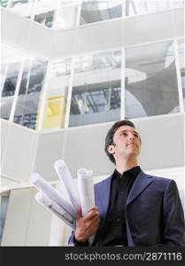 Business man holding rolled blueprints under arm in atrium of office building low angle view