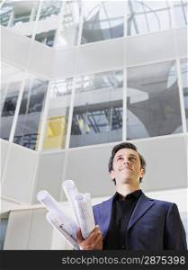 Business man holding rolled blueprints under arm in atrium of office building low angle view
