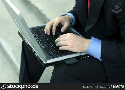 Business man in black suit, blue shirt, and red tie is busily typing on laptop on courthouse steps