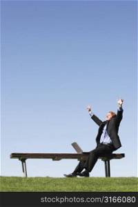 Business man raises arms in triumphant victory as he sits on a park bench, next to his laptop against the blue sky and green grass