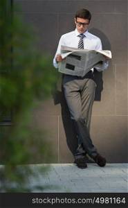 Business man reading a newspaper on the street leaning on wall of modern building