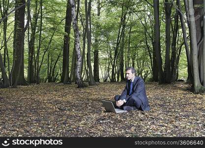 Business man sitting on ground using laptop in middle of forest