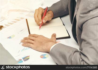 business man sitting writing text note black cover on graph diagram paper, he wear gray suit and use red pen on work table at office