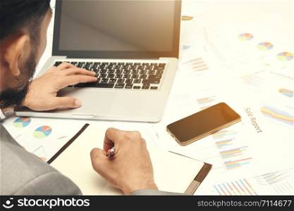 business man sitting writing text note black cover on graph diagram paper looking computer laptop, he wear gray suit and use red pen and smartphone on work table at office