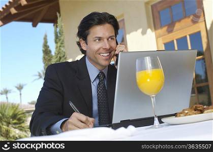 Business man talking on phone and writing in front of laptop, outdoors