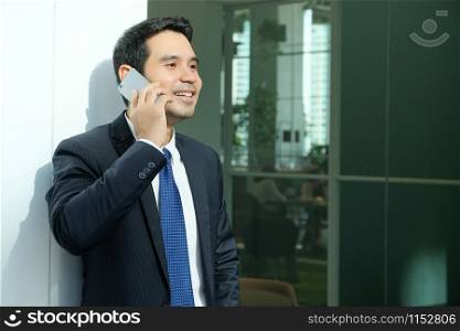 Business man talking smart phone with happy smiling face standing in front of meeting room inside office building background, businessman on smart phone, online conferance concept