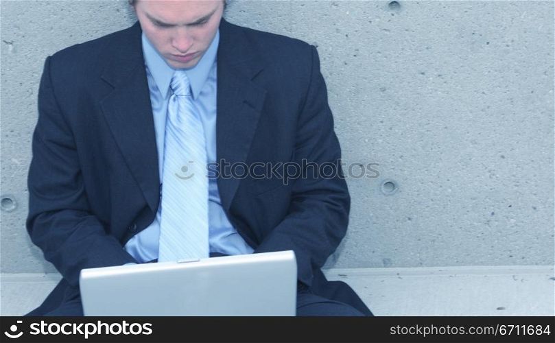 Business man with blue suit, blue shirt, and blue tie is sitting up against a concrete wall typing on his laptop