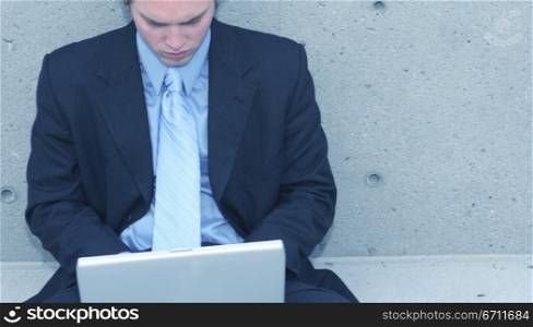 Business man with blue suit, blue shirt, and blue tie is sitting up against a concrete wall typing on his laptop