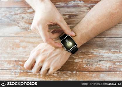 business, modern technology, startup, idea and people concept - close up of male hands setting smart watch with light bulb on screen on wooden table
