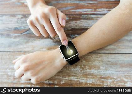 business, modern technology, startup, idea and people concept - close up of female hands setting smart watch with light bulb on screen on wooden table