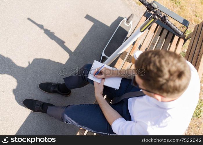 business news and corporate people and concept - young businessman with bag, electric scooter and coffee sitting on street bench and writing to notebook in city. smiling businessman writing to notebook in city