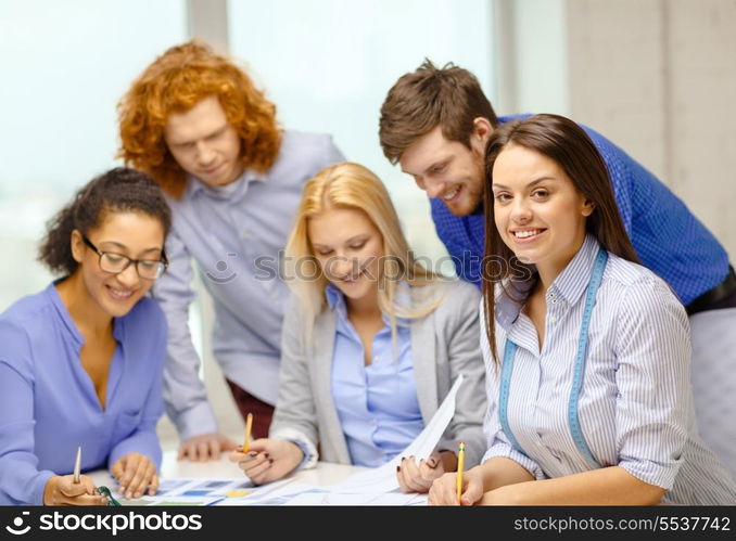 business, office, clothes design and starup concept - smiling female tailor with team on the back looking over clothes designs