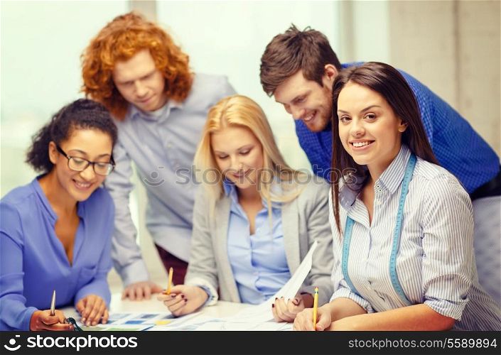 business, office, clothes design and starup concept - smiling female tailor with team on the back looking over clothes designs