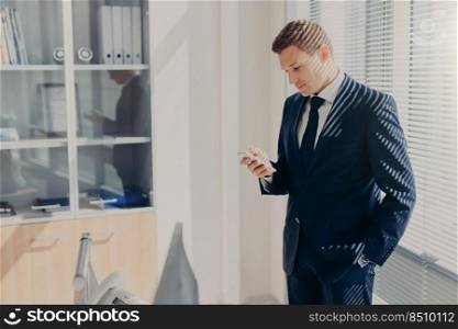 Business, office, people and technology concept. Successful businessman holds smart phone, reads text message from investor, stands in his cabinet, wears formal clothes, connected to wifi at office