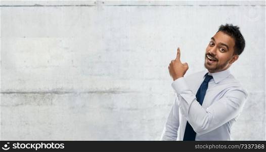 business, office worker and people concept - smiling indian businessman in shirt with tie pointing finger at something invisible over gray concrete wall background. indian businessman pointing finger at wall
