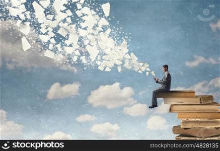 Business online. Young businessman sitting on pile of books with mobile phone in hands