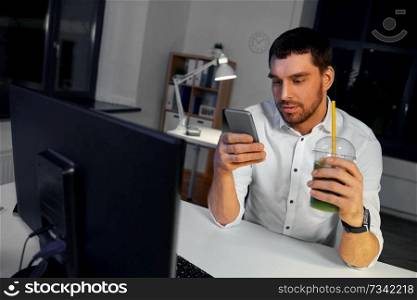 business, overwork, deadline and people concept - businessman using smartphone and drinking green smoothie at night office. businessman with drink using smartphone at office