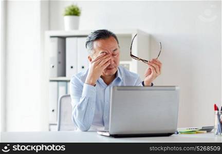 business, overwork, deadline and people concept - tired businessman with eyeglasses and laptop computer rubbing eyes at office. tired businessman with glasses at laptop in office