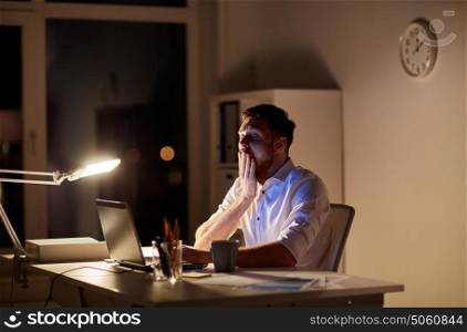 business, overwork, deadline and people concept - tired man with laptop working at night office and yawning. man with laptop and coffee working at night office