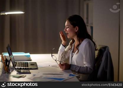 business, overwork, deadline and people concept - tired woman with eyeglasses and laptop yawning at night office. tired woman with laptop yawning at night office