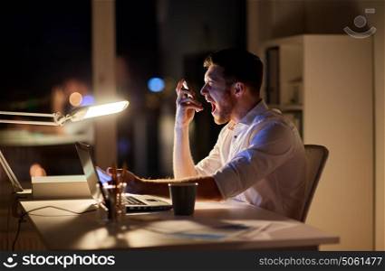 business, overwork, people, deadline and technology concept - angry businessman with laptop computer calling on smartphone at night office and screaming. angry businessman with smartphone at night office