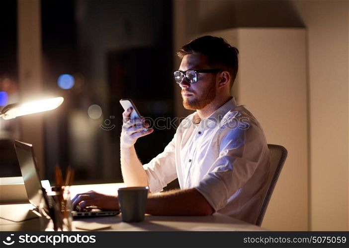 business, overwork, people, deadline and technology concept - businessman in glasses with laptop computer texting on smartphone at night office. businessman texting on smartphone at night office
