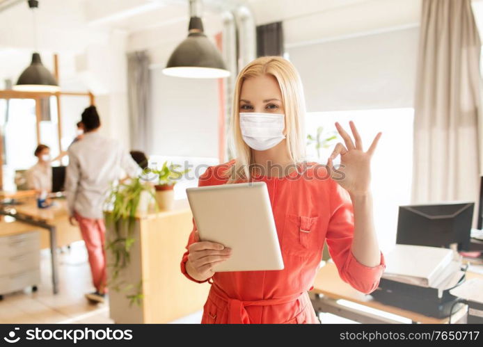 business, pandemic and health concept - businesswoman or creative female office worker in face protective medical mask for protection from virus disease with tablet pc computer showing ok hand sign. woman in mask with tablet pc showing ok at office
