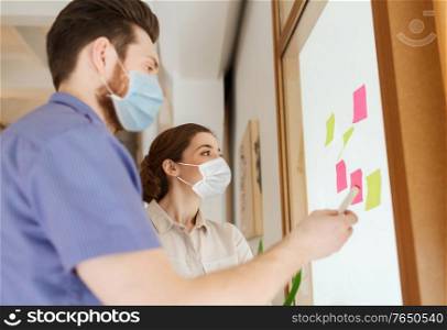 business, pandemic and health concept - creative team wearing face protective medical masks for protection from virus disease with sticky notes on office glass board. team in masks with stickers on glass at office