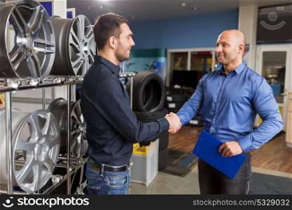 business, partnership and people concept - male customer and salesman shaking hands at car repair service or auto store. auto business owner and wheel rims at car service