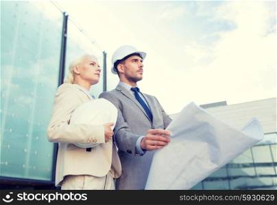 business, partnership, architecture and people concept - businessman and businesswoman with blueprint and helmets on city street