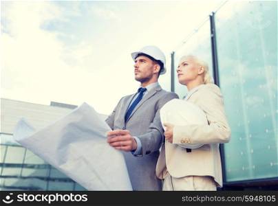 business, partnership, architecture and people concept - businessman and businesswoman with blueprint and helmets on city street