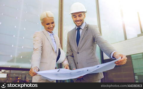 business, partnership, architecture and people concept - smiling businessman and businesswoman with blueprint and helmets on city street