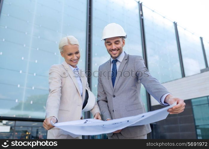 business, partnership, architecture and people concept - smiling businessman and businesswoman with blueprint and helmets on city street