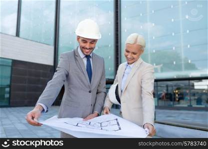 business, partnership, architecture and people concept - smiling businessman and businesswoman with blueprint and helmets on city street