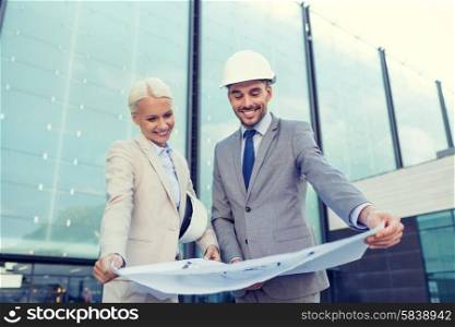 business, partnership, architecture and people concept - smiling businessman and businesswoman with blueprint and helmets on city street