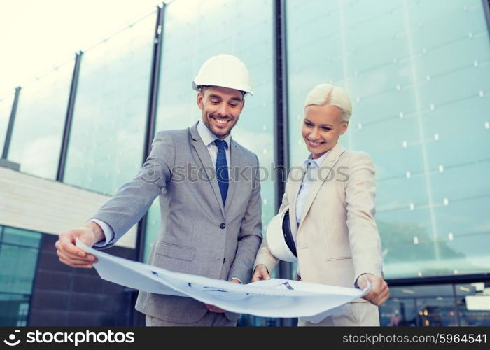 business, partnership, architecture and people concept - smiling businessman and businesswoman with blueprint and helmets on city street