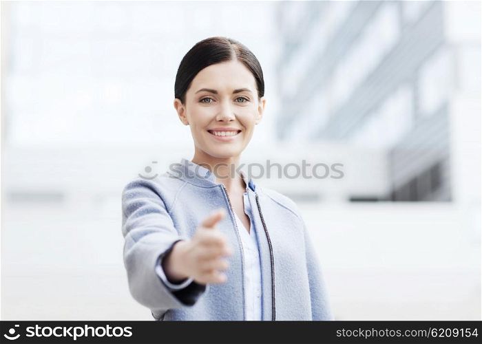 business, partnership, cooperation and people concept - young smiling businesswoman giving hand for handshake over office building