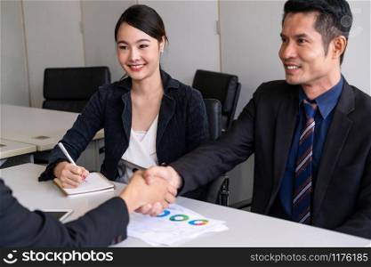Business people agreement concept. Asian Businessman do handshake with another businessman in the office meeting room. Young Asian secretary lady sits beside him.