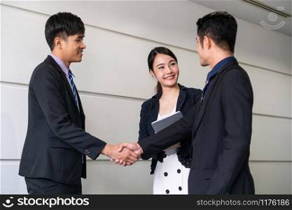 Business people agreement concept. Businessman do handshake with another businessman in the office meeting room. Young Asian secretary lady stands beside them.