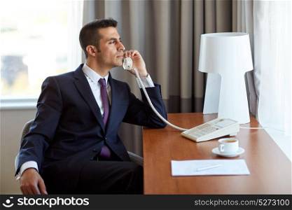 business, people and communication concept - businessman with coffee and papers calling on desk phone at hotel room. businessman calling on desk phone at hotel room
