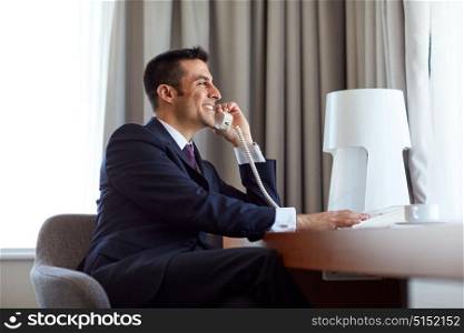 business, people and communication concept - happy smiling businessman with coffee calling on desk phone at hotel room. businessman calling on desk phone at hotel room