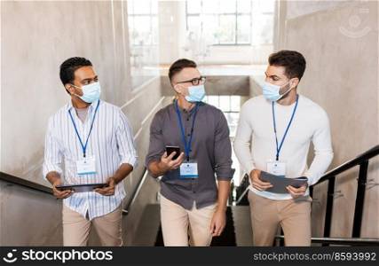 business, people and corporate concept - businessmen in protective medical masks with name tags walking up office stairs and talking. businessmen in masks with name tags going upstairs