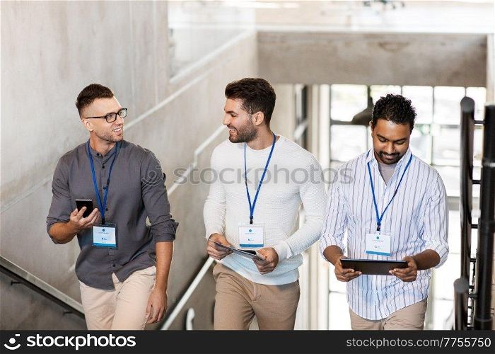 business, people and corporate concept - group of men with conference badges, folder, tablet pc computer and smartphone walking up office stairs. men with conference badges walking upstairs
