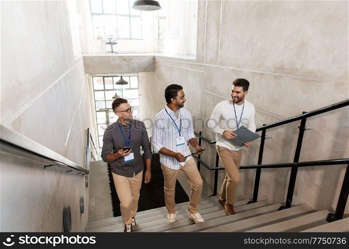 business, people and corporate concept - men with conference badges, folder, tablet pc computer and smartphone walking up office stairs. men with conference badges walking upstairs