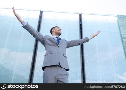 business, people and education concept - young smiling businessman with raised hands over office building