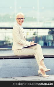 business, people and education concept - young smiling businesswoman in glasses with notepad over office building