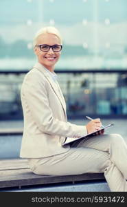 business, people and education concept - young smiling businesswoman in glasses with notepad over office building