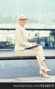 business, people and education concept - young smiling businesswoman in glasses with notepad over office building