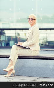 business, people and education concept - young smiling businesswoman in glasses with notepad over office building